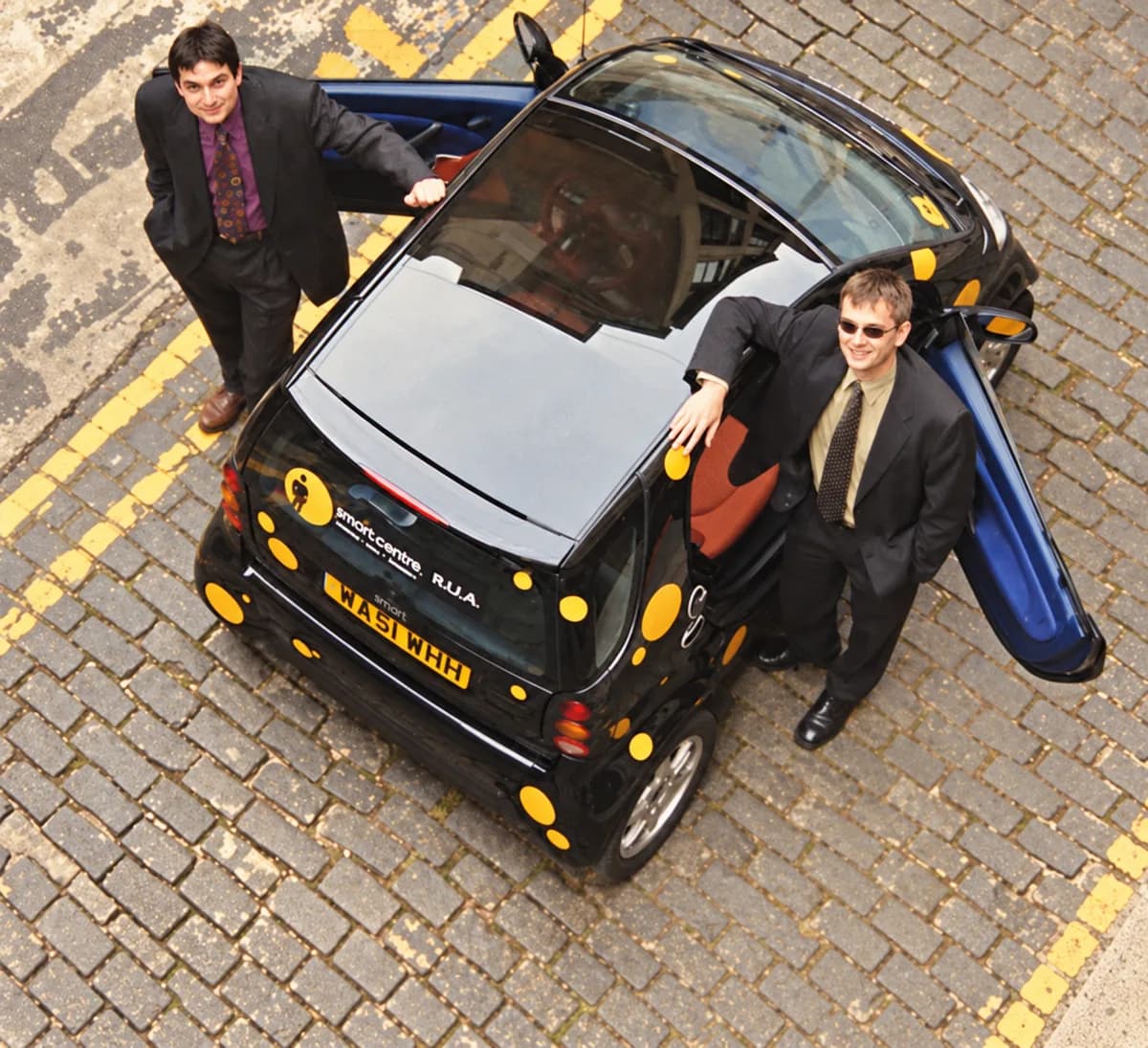 Two members of the On the Spot team, photographed from above, standing beside a black smart car covered in yellow spots.